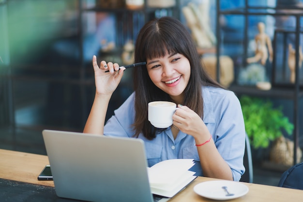 Азиатская красивая женщина в синей рубашке, используя ноутбук и пить кофе