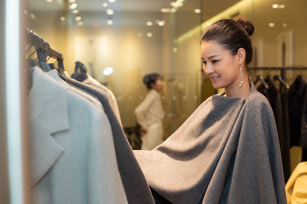 asian beautiful woman in dress select new collection on gray earth tone clothes rack in retail fashion store which just open brand news for winter autumn as minimal style