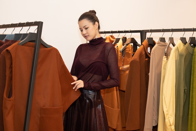 Asian Beautiful Woman in dress select new collection on orange green earth tone clothes rack in retail fashion store which just open brand news for winter autumn as minimal style