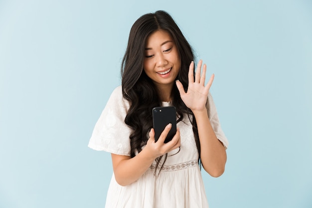 Asian beautiful woman isolated over blue wall talking by mobile phone waving