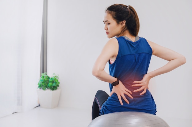 Bella donna asiatica che si siede sul fitball e che sente mal di schiena mentre gioca yoga ed eserciti a casa.