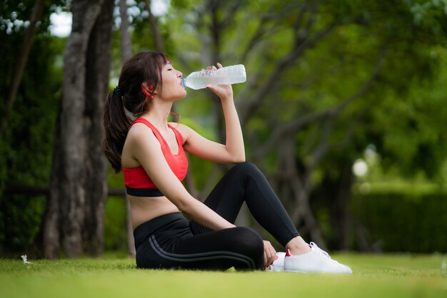 アジアの美しい女性は、一人でトレーニングの後、村の公園でボトルから水を飲んでリラックスします。