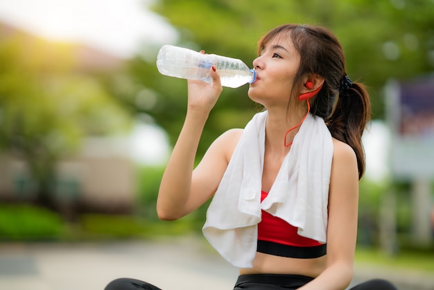 La bella donna asiatica che si siede per si rilassa dopo l'allenamento da solo e l'acqua potabile da imbottiglia il parco pubblico in villaggio