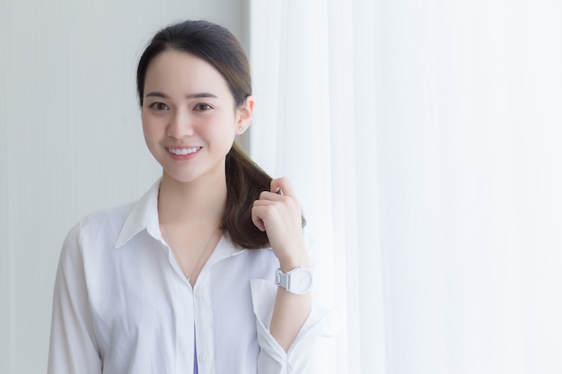 La bella donna asiatica in camicia bianca sta sorridendo e stando vicino alla finestra con la tenda bianca.