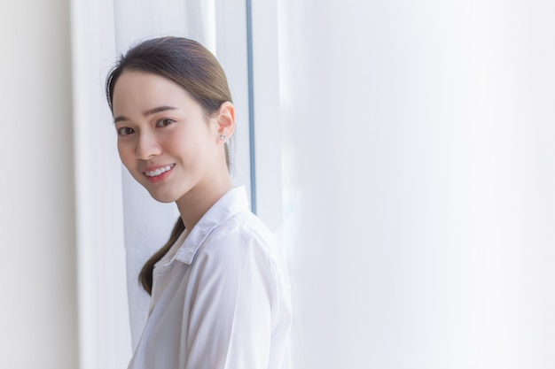 La bella donna asiatica in camicia bianca sta sorridendo e stando vicino alla finestra con la tenda bianca.