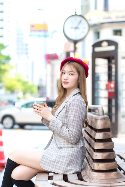 Asian beautiful woman who wears suit and red cap with bronze hair sits on chair in the city outdoors