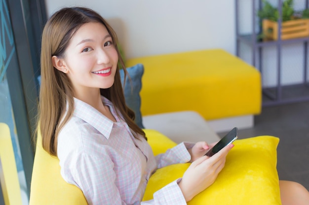 셔츠를 입은 긴 머리를 가진 아시아의 아름다운 여성이 스마트폰을 하는 동안 노란 소파에 앉아 있다