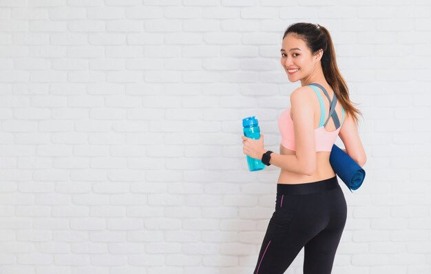 Le belle donne asiatiche che tengono la bottiglia di acqua e l'yoga stuoia dopo yoga del gioco ed esercitano su bianco