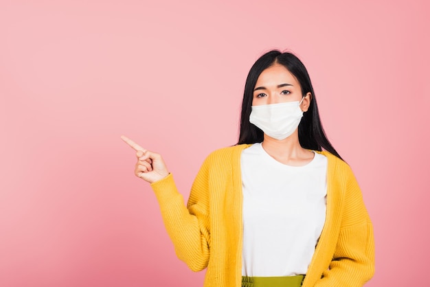 Asian beautiful young woman wearing medical mask protection prevent infection coronavirus, COVID-19 she's pointing finger to space, studio shot isolated on pink background, medical health care concept