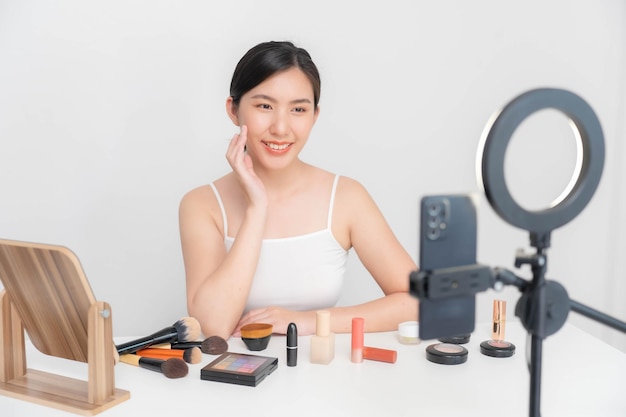 Asian beauty blogger doing makeup tutorial on mobile phone at home using white wall backdrop