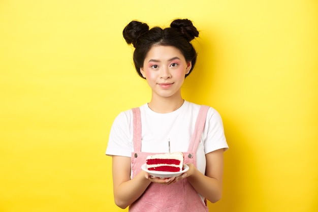 Asian birthday girl standing with cake and smiling, celebrating b-day on yellow.