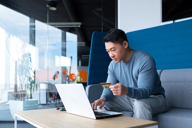 Asian boss transfers money from a bank card, sitting in the office, satisfied with the result businessman smiles, works on laptop