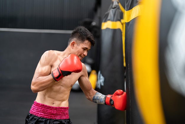 Foto uomo atleta pugile asiatico con guantoni da boxe rossi che si allena punzonando il sacco di sabbia in palestra