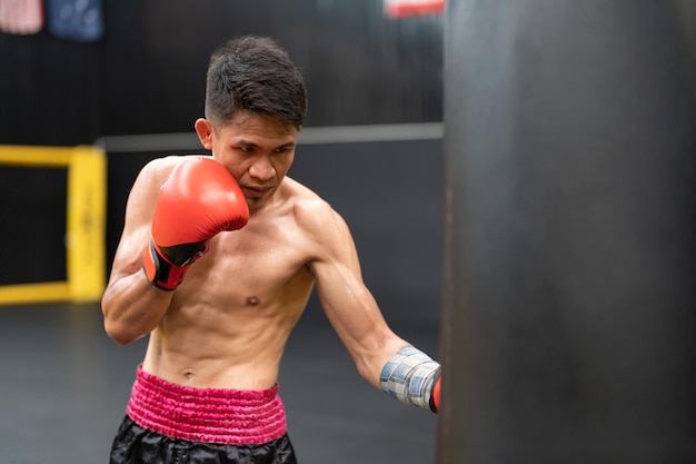 Foto uomo atleta pugile asiatico con guantoni da boxe rossi che si allena punzonando il sacco di sabbia in palestra