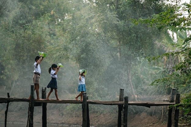 학생복을 입은 아시아 소년과 어린 소녀 두 명, 맨발로 머리 위에 바나나 잎을 들고, 아침에 학교에 가는 나무 다리를 걷고, 복사 공간, 시골 풍경 스타일 개념