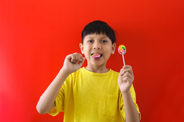 アジアの少年は赤い背景にロリポップキャンディーをかみます