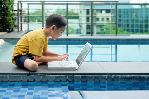 Asian boy child sitting side of pool and uses laptop for education