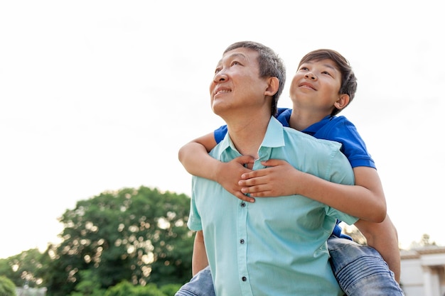 아시안 소년이 여름에 아빠의 등 위에서 앞으로 날아다니며 한국의 고령 연금자는 아이와 놀고 있습니다.