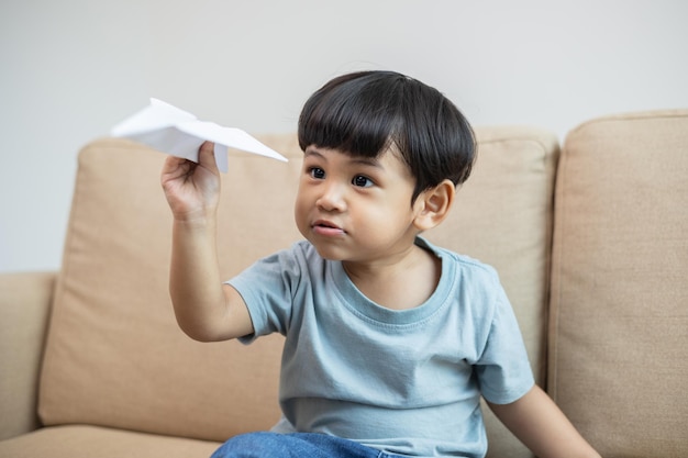 Ragazzo asiatico piega un aeroplano di carta per volare gioca a casa nel soggiorno sviluppare l'apprendimento dei bambini prima di entrare nella scuola materna pratica le abilità