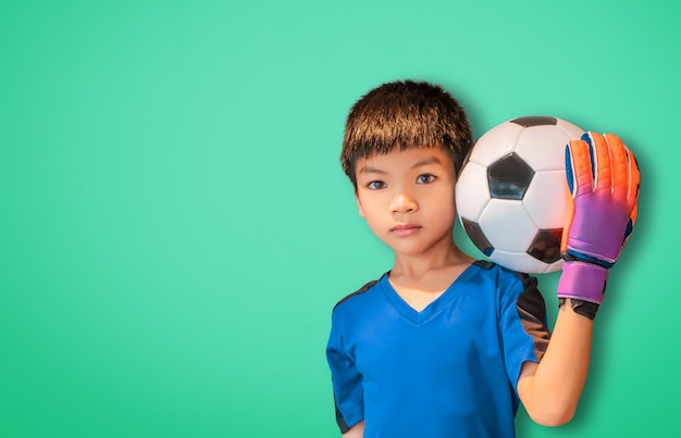 아시아 소년은 장갑을 끼고 녹색 배경 복사 공간에 축구공을 들고 있는 축구 골키퍼입니다.