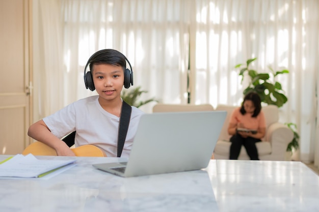 Ragazzo asiatico che suona la chitarra e guarda il corso online sul laptop mentre si esercita per imparare la musica
