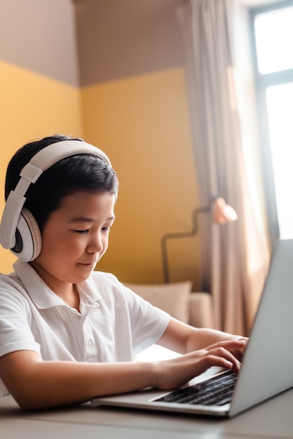 検疫中に自宅でラップトップとヘッドフォンを使ってオンラインで勉強しているアジアの少年