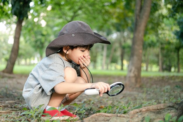사진 숲 탐사 의복 을 입은 모자를 입은 아시아 소년 은 나무 지역 을 조사 하기 위해 확대 유리 를 사용 한다