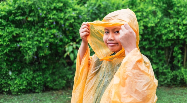 オレンジ色のレインコートを着たアジアの少年は幸せで、雨の日の雨の中で楽しんでいます