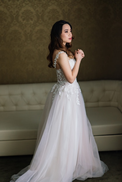 Asian bride in wedding dress in hotel sitting on the bed, standing near window, morning