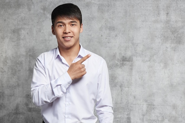 Asian in business clothes advertises something. Young Kazakh with joyful facial expression points finger to the side on free space for your product.