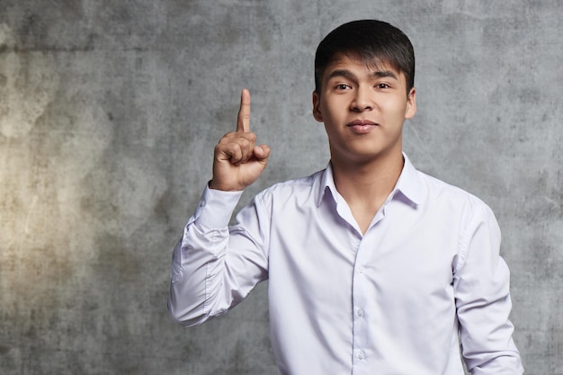 Asian in business clothes advertises something. Young Kazakh with joyful facial expression points finger up on free space for your product.