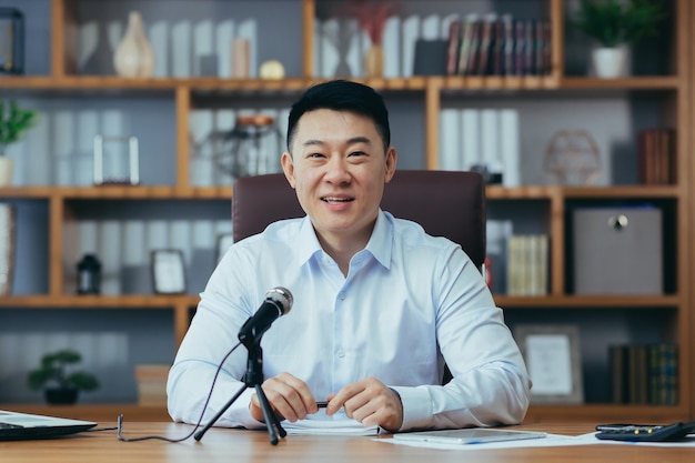 Азиатский бизнес-тренер записывает аудиоподкаст урок экономики бизнесмен за столом использует микрофон смотрит в камеру и улыбается портрет
