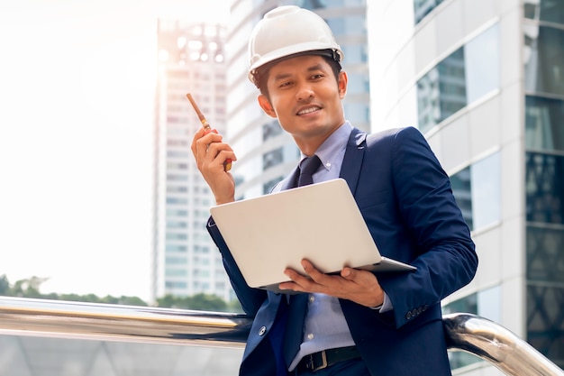 Uomo asiatico del responsabile dell'ingegnere di affari che sta e che lavora con il computer portatile alla città all'aperto.