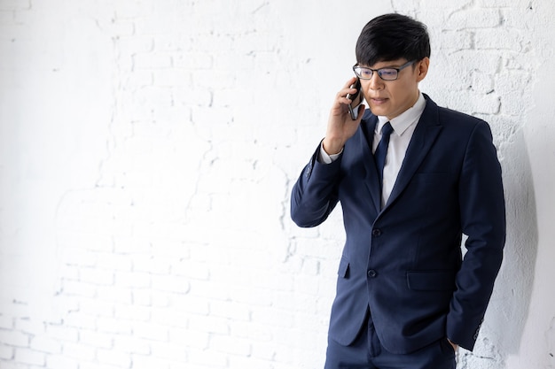 Asian business in eyeglasses stands on white wall background uses smart phone for making calls business, Businessman working by using smart phone.