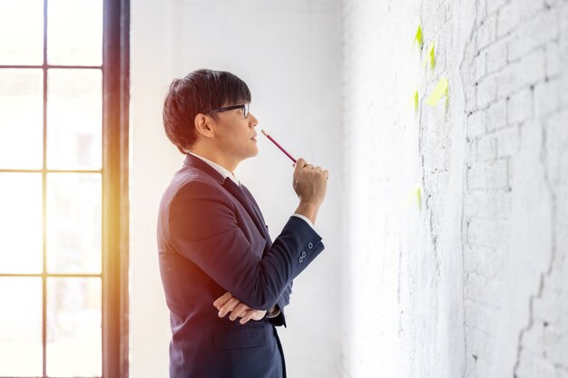 Asian business in eyeglasses stands on window use sticky notes paper reminder schedule for discussing idea in office, businessman looking and thinking notes pad in the white wall.