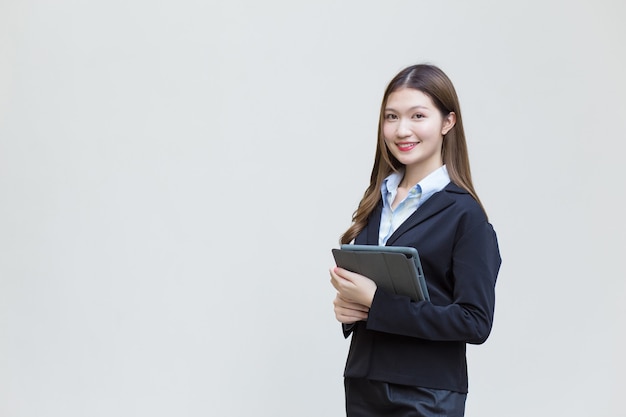 Asian business female in black suit smiles happily while she works and holds tablet on white background.