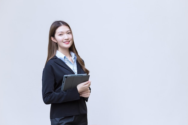 Asian business female in black suit smiles while she works and holds tablet on white background