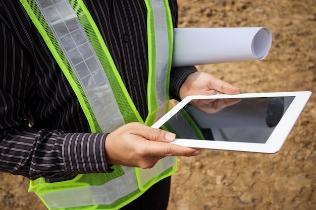 Asian business man construction manager and engineer worker in protective helmet hold blueprints paper on hand using tablet computer at building site