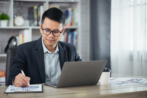 Asian business man smiling using pen making notices in notebook and working on laptop on wooden table at home office Entrepreneur man working for business at home Business work at home concept