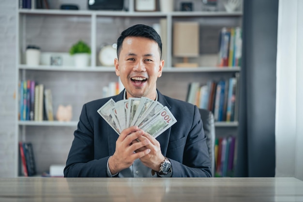 아시아 사업가는 달러 지폐를 들고 웃으면서 집 사무실 거실에 앉아 있는 책상에 앉아 돈을 벌고 있다