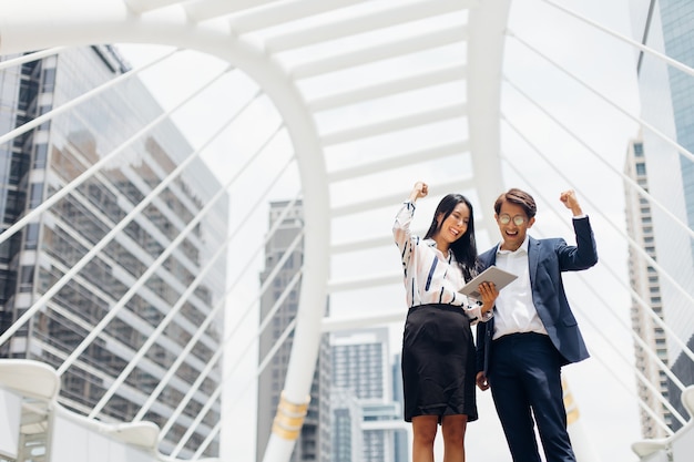 Asian business man talking on mobile phone and young beautiful woman working on digital tablet computer standing outside office building.