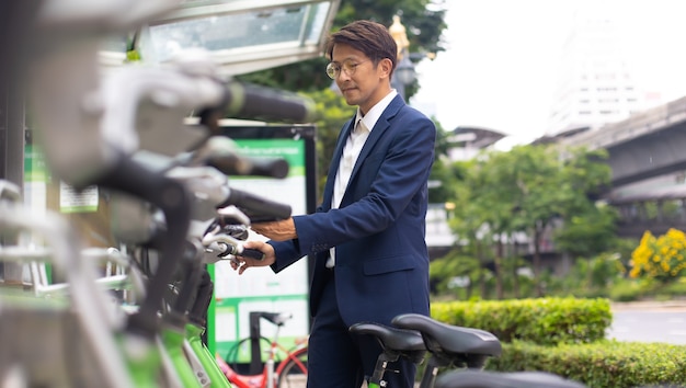 屋外で公共の自転車を借りるためにスマートフォンを使用しているアジアのビジネスマン。都市環境でのレンタル自転車。