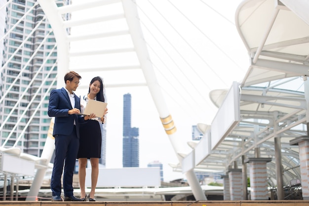 Asian business man and woman working on laptop computer standing outside office building.