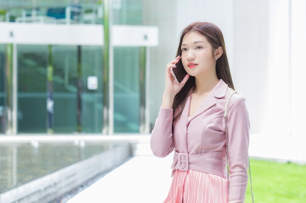 Asian business professional in pink dress is calling phone seriously with someone at a building