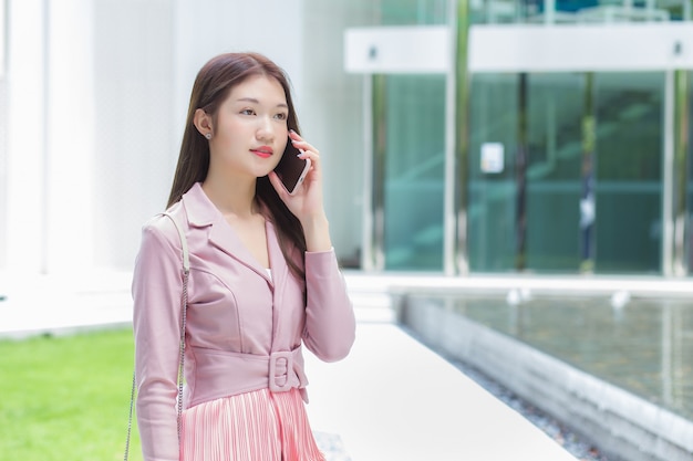 ピンクのドレスを着たアジアのビジネス専門家の女性は、建物の誰かと真剣に電話をかけています