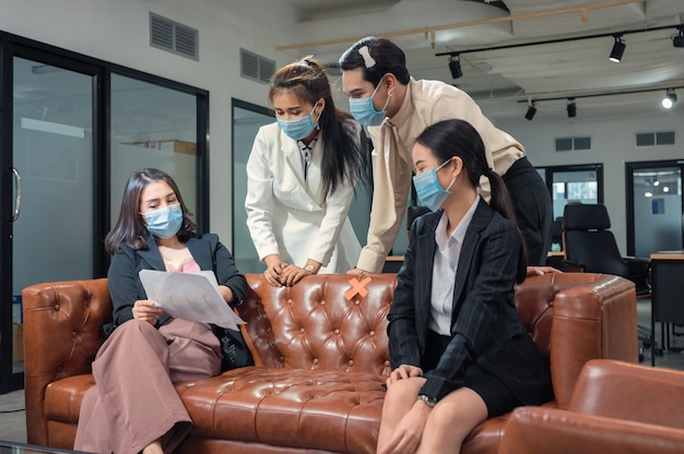 Азиатская бизнес-команда в маске обсуждает бизнес-план на кожаном диване в новом обычном офисе