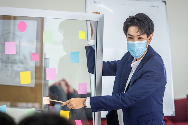 Asian Business tomboy lesbian wearing protective mask presenting use post it notes to share idea.Brainstorming concept.Sticky note on glass wall in the meeting room at office,Concept of COVID-19