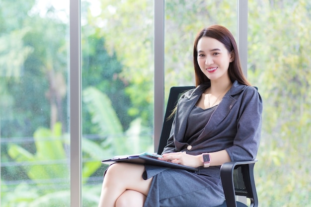 Asian business woman in dark grey dress  sits on chair in office room and works happily.