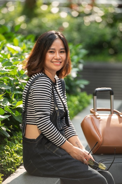 Asian Business Woman with yellow glasses sitting in the park with her suitcase in the background