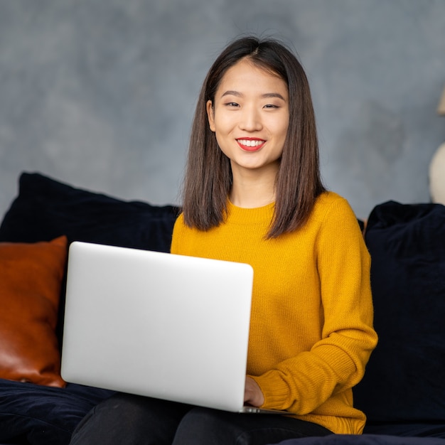 Asian business woman working in laptop on sofa. Japanese businesswoman work at home. Active serious smart female worker looking at camera